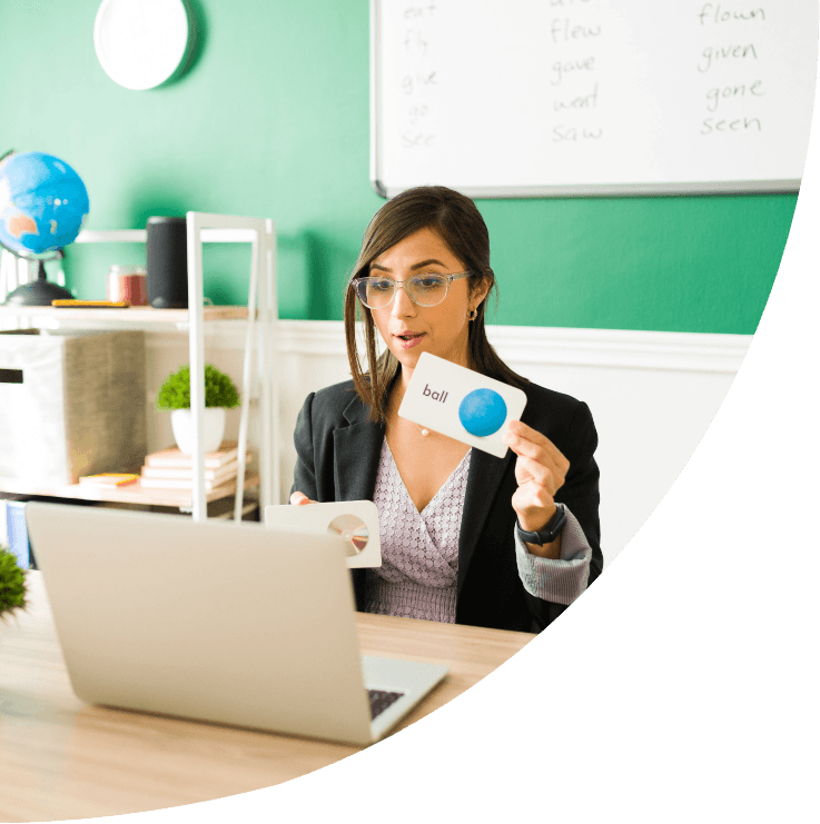 Fotografia de uma professora de óculos ministrando uma aula de inglês online. Sentada à frente de um laptop, ela segura um cartão educativo com a palavra ball e a imagem de uma bola azul.